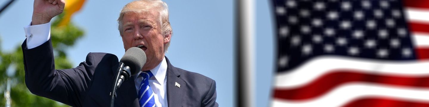 Trump che fa un comizio, sul palco, con il microfono in mano e la bandiera americana come sfondo