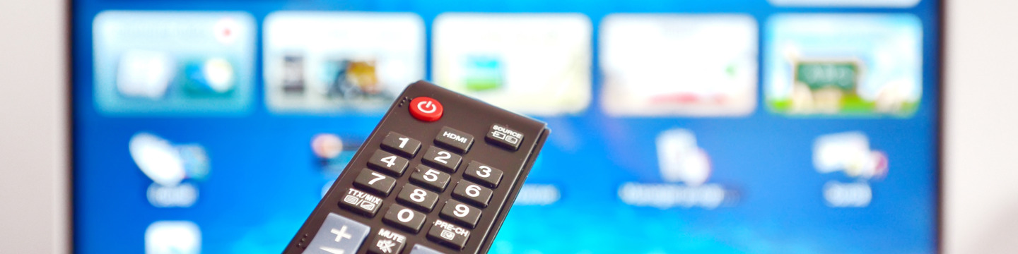 Tv_websim2 Telecomando in primo piano con pulsanti visibili; sullo sfondo si intravede una TV accesa sfocata.