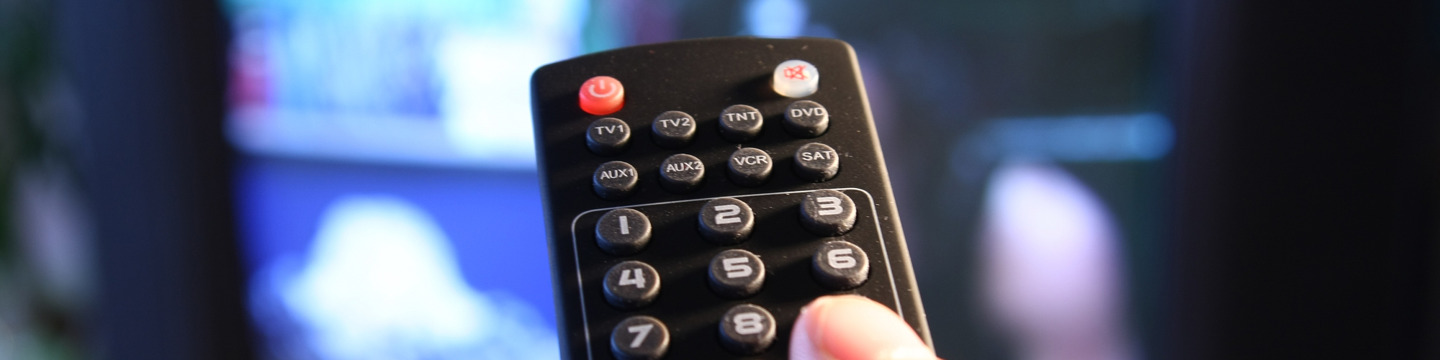 Dettaglio di una mano che punta il telecomando verso una TV accesa, pronta a cambiare canale.