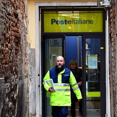 Ingresso di un ufficio postale Poste Italiane, con logo giallo-blu e persone in entrata e uscita