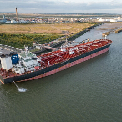 Profilo laterale di una grande nave cargo, con container impilati sul ponte e mare calmo intorno.