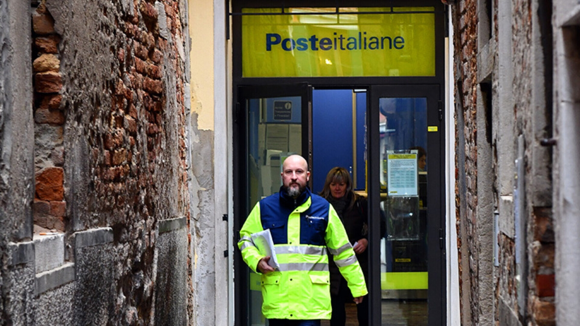 Ingresso di un ufficio postale Poste Italiane, con logo giallo-blu e persone in entrata e uscita
