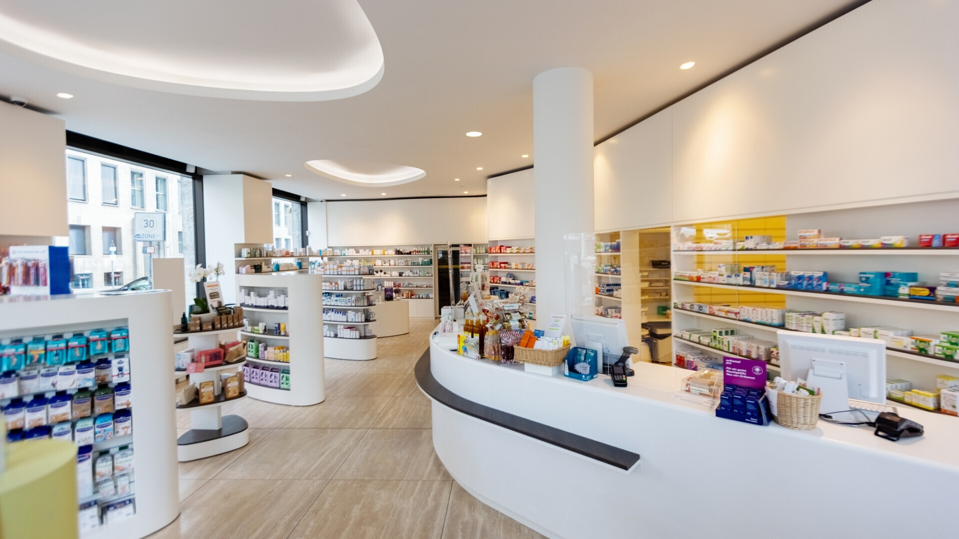 Interno di una farmacia moderna, con scaffali e ripiani pieni di medicinali, senza persone presenti.